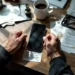 Business owner's hands gripping desk edge with smartphone showing marketplace app, rejection letters scattered below with spilled coffee