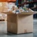 Kartonnen verzenddoos gevuld met diverse verpakte producten op een magazijnvloer, met pallets en hoge stellingen op de achtergrond.