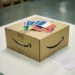 Amazon shipping box on warehouse desk with European country flags reflected on surface, VAT registration document visible underneath.