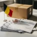 Belgian tax documents and invoices fanned on a white desk beside a small Belgian flag and sealed shipping box in an e-commerce office.