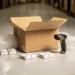 Open cardboard shipping box on warehouse floor surrounded by bubble wrap, barcode scanner, and packaged products ready for dispatch.