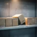 Dust-covered product boxes on a dim warehouse shelf, one tipped on its side, with concrete walls and warm overhead light casting long shadows.