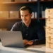 Entrepreneur reviewing product listings on a laptop beside stacked branded shipping boxes, with a softly blurred warehouse shelf in warm golden-hour light.