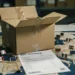 Open cardboard shipping box on a warehouse desk with EU country flag pins on a paper map of Europe and a partial VAT invoice underneath.