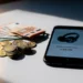 Euro coins and folded banknotes beside a smartphone showing an Amazon product listing on a white desk.