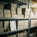 Stacked cardboard boxes on industrial metal warehouse shelves with a clipboard and measuring tape resting against the bottom shelf.