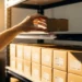 Hand placing a cardboard box onto a warehouse shelf stocked with identical product boxes, warm golden light casting soft shadows.
