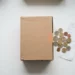 Cardboard shipping box on a white desk beside scattered euro coins and a folded receipt, warm beige tones, flat lay composition.
