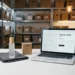 Skincare bottle, folded garment, and electronics box arranged on white surface beside laptop displaying a product listing template, e-commerce warehouse shelving in background.