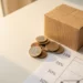 Stack of euro coins next to a small cardboard shipping box on a white desk with a percentage breakdown sheet beneath, warm golden light.