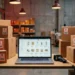 Laptop displaying an e-commerce product listing in a warehouse surrounded by cardboard boxes with red price tags and a barcode scanner.