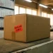 Sealed cardboard shipping box with a red Kaufland price tag on a warehouse conveyor belt, wall calendar softly blurred in the background.