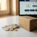 Polish złoty coins and banknotes stacked beside a small cardboard shipping box on a white desk, laptop blurred in background.