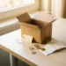 Open cardboard shipping box on a white desk with scattered euro coins, folded receipts, and a price tag in warm natural light.