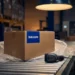 Verzegelde kartonnen verzenddoos op magazijnband met blauw etiket, borrelverpakking en barcodescanner op de voorgrond.