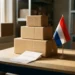 Stapel kartonnen verzenddozen op een magazijntafel met een factuur en een kleine Nederlandse vlag in warm zijlicht.