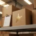 Stacked cardboard shipping boxes on a warehouse shelf, one featuring a gold star rating badge, beside a quality checklist clipboard.